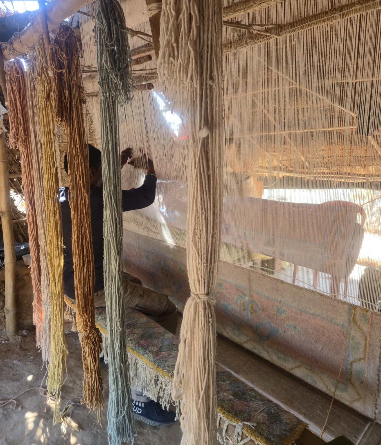 An artisan hand-knotting a rug at a vertical loom, surrounded by yarn.